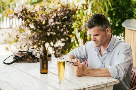 Man with a camera having a relaxing break at the bar, he is using a digital tablet and drinking a beerの写真素材