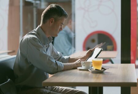 Businessman having breakfast at the bar, he is sitting next to a window and working with a digital touch screen tabletの写真素材