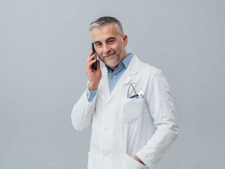 Confident mature doctor having a phone call with his smartphone: medical service consultation by phoneの写真素材