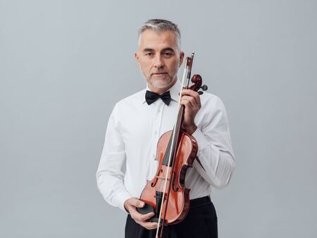 Confident mature musician posing with a violin and looking at camera, classical music and entertainment conceptの写真素材