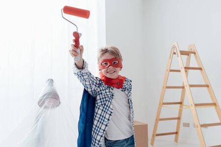 Cute superhero boy with mask and cape, he is holding a paint roller and smiling at camera, home renovation and diy conceptの写真素材