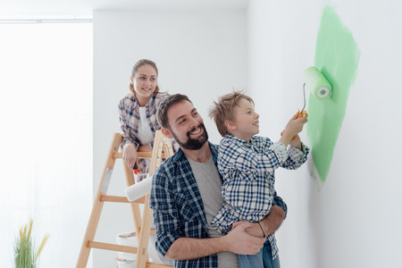 Happy young family renovating their home, the father is holding his son and he is helping him to paint a wall with a paint roller, the mother is standing on the ladder and smilingの写真素材