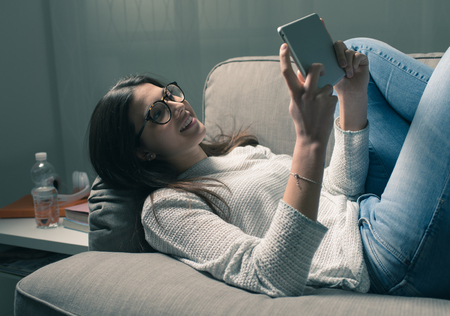 Smiling young girl relaxing on the sofa late at night and connecting with a digital touch screen tabletの写真素材