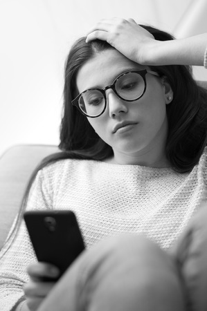 Sad young girl lying on the sofa and texting with her smartphone, she is sad and pensiveの写真素材