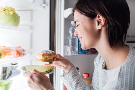 Woman having an unhealthy snack, she is taking a delicious pastry out of the fridgeの写真素材