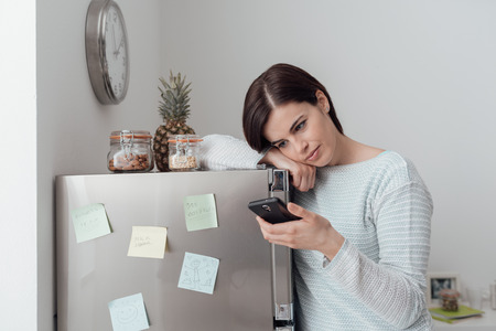 Smiling woman relaxing in the kitchen and texting with her mobile phone, she is leaning on the fridgeの写真素材