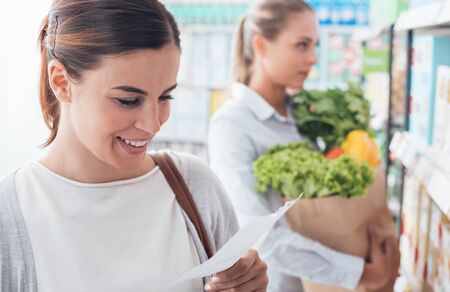 Women shopping at the supermarket, they are holding a grocery bag with fresh vegetables and checking a listの写真素材