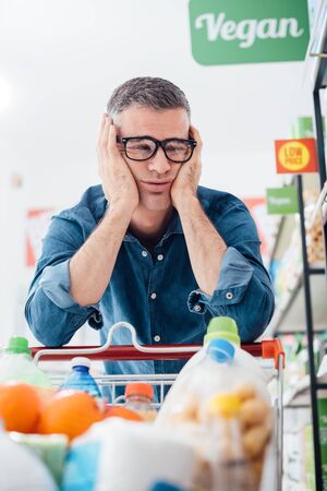 Sad exhausted man shopping at the supermarket, he is leaning on the full shopping cart with head in handsの写真素材
