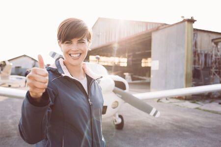 Smiling young pilot posing at the airport and giving a thumbs up, propeller plane on the background, travel and aviation conceptの写真素材