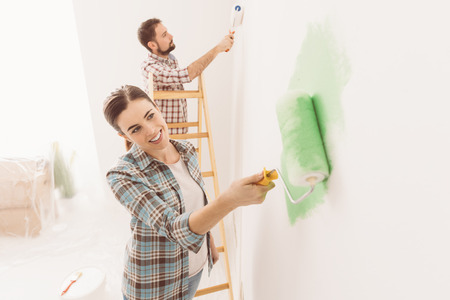 Happy couple painting walls in their new house: the man is standing on a ladder and the woman is using a paint roller and applying bright green paintの写真素材