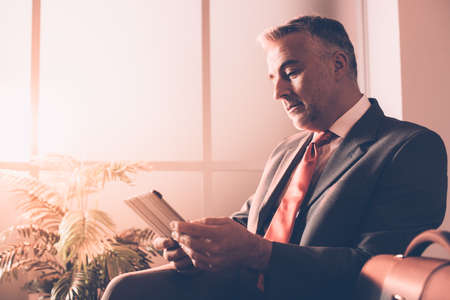 Corporate businessman connecting with a digital touch screen tablet, he is sitting in the waiting roomの写真素材