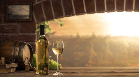White wine tasting in the wine cellar: wineglass and bottles next to the window and panoramic view of vineyards at sunsetの写真素材