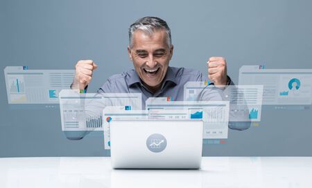 Cheerful successful businessman reading positive financial reports on visual screens and working with a laptopの写真素材