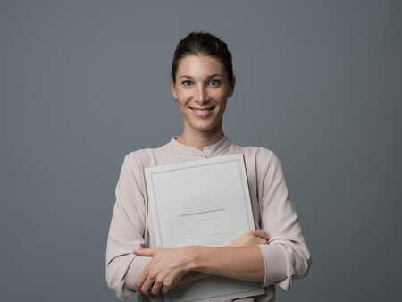 Professional female secretary holding paperwork and smiling at cameraの写真素材