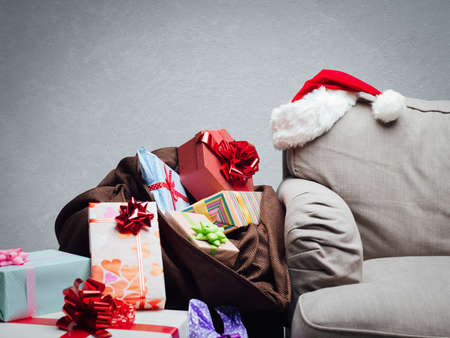 Load of colorful gifts in a sack next to a sofa in the living room and Santa's hat: Christmas celebration at home conceptの写真素材
