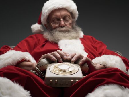 Santa Claus falling asleep while waiting for a phone call, he is relaxing on the armchair and holding a vintage phone on his lapの写真素材