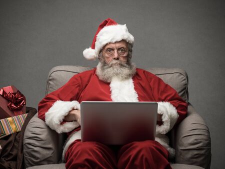 Santa claus relaxing at home and connecting with a laptop, he is chatting and social networking, Christmas time and technology conceptの写真素材