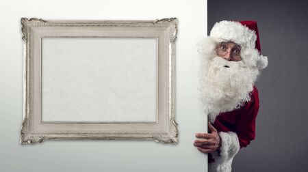 Santa Claus peeking behind a wall and blank decorative frame, Christmas and holidays conceptの写真素材