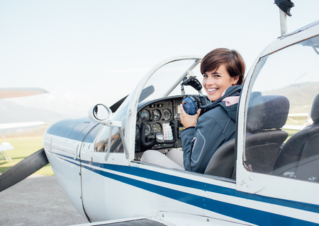Smiling female pilot in the light aircraft cockpit, she is holding aviator headset and looking at cameraの写真素材