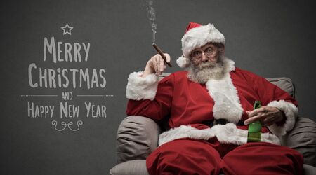 Bad Santa celebrating Christmas at home alone, he is smoking a cigar and drinking beer, Christmas card with wishesの写真素材