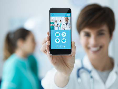 Smiling confident female doctor holding a touch screen smartphone and medical staff on the background, medical app conceptの写真素材