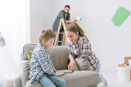 Happy young family renovating and decorating their home, the boy is picking colors swatches with his mother and his father is painting on the backgroundの写真素材