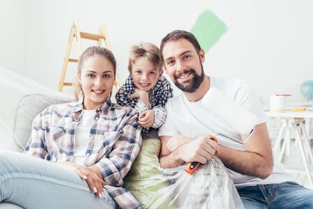 Happy family renovating their home and painting rooms, they are sitting together and smiling, the father is holding a paint rollerの写真素材
