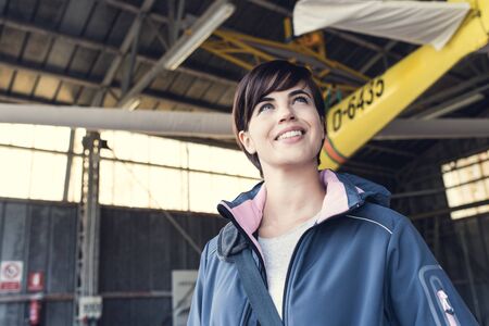 Smiling female pilot posing in the airport hangar, propeller airplane on the background, travel and aviation conceptの写真素材