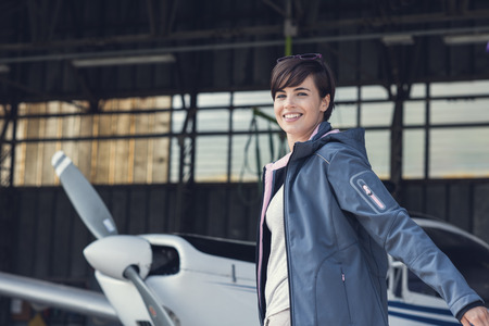Smiling cheerful female pilot posing in the hangar with arms raised like wings, propeller plane on the background, travel and aviation conceptの写真素材
