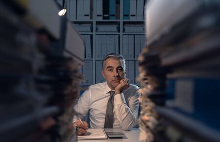 Stressed exhausted business executive working in the office late at night with piles of paperwork, he is overloaded with work: management and deadlines conceptの写真素材