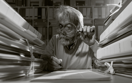 Senior researcher using a magnifying glass and writing, he is surrounded by stacks of paperworkの写真素材