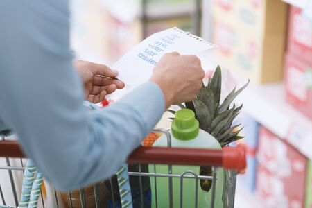 Woman doing grocery shopping at the supermarket, she is pushing a full shopping cart and checking a...