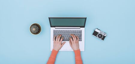 Minimalist light blue workspace with laptop, digital camera and cactus, a woman is typing on the keyboard, top viewの写真素材