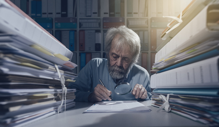 Senior researcher using a magnifying glass and writing, his desk is coverer with stacks of paperworkの写真素材