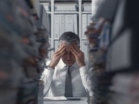 Stressed exhausted business executive in the office overloaded with work, he has piles of paperwork on the deskの写真素材