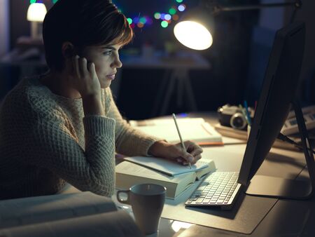Young woman working and studying late at night, she is connecting with her computer and writing down...