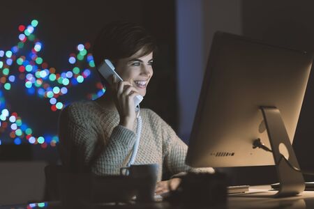 Young smiling woman working with her computer and talking on the phone late at night, she is holding...