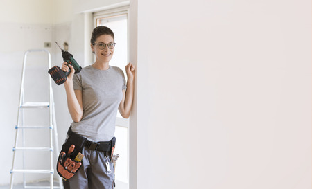 Professional repairwoman with tool belt doing a home renovation, she is posing and holding a drill