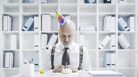 Senior businessman having a sad lonely birthday in the office, he is wearing a party hat and having a small cake aloneの写真素材