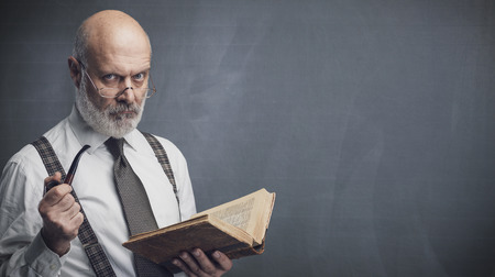 Confident senior professor smoking a pipe and reading an old book, knowledge and education conceptの写真素材