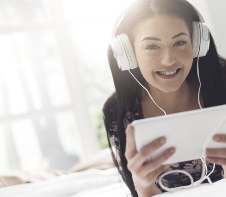 Happy young woman lying in bed and watching videos online with her tablet, she is wearing headphonesの写真素材