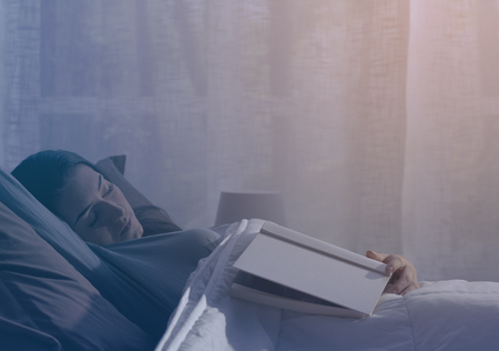 Young woman relaxing in her bed at night, she is falling asleep while reading a bookの写真素材
