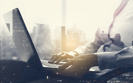 Corporate American businesswoman sitting next to a window and connecting with her laptop on her lap, business and communication concept, city skyline in the backgroundの写真素材