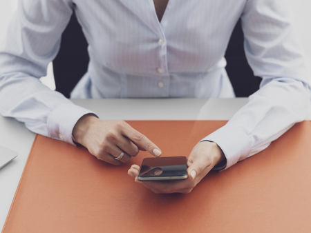 Professional corporate businesswoman sitting at office desk and working, she is connecting with her smartphone, business and communication conceptの写真素材