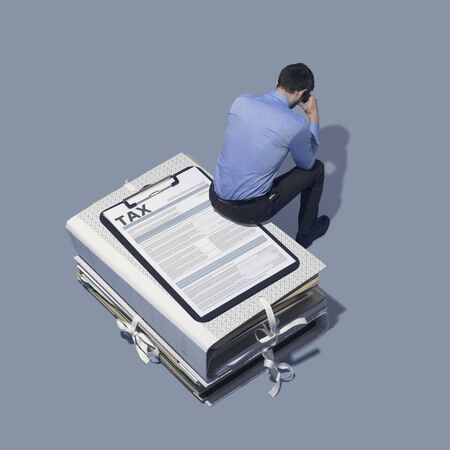 Stressed businessman sitting on a tax form and a pile of paperwork, deadlines and management conceptの写真素材