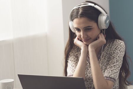 Happy smiling woman wearing headphones and watching movies online with her laptop, entertainment and...