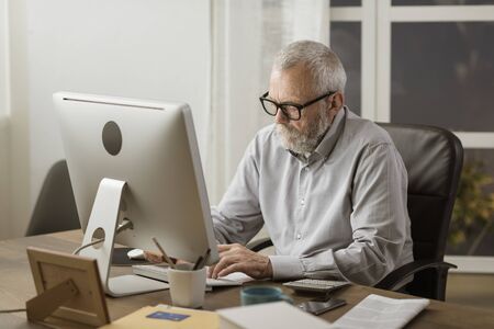 Senior retired man sitting at desk and working with a computer, elderly and technology conceptの写真素材