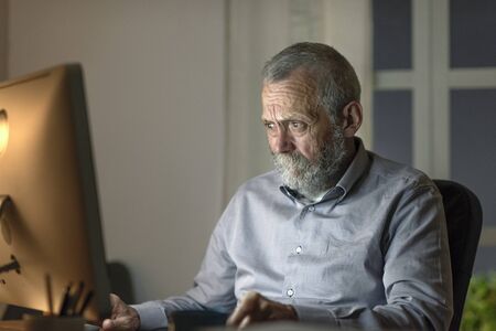 Senior man sitting at desk and working with his computer late at night, he is staring worried at the...