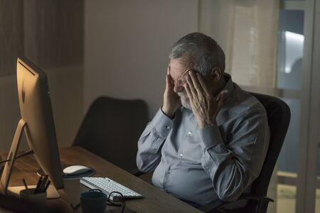Lonely sad senior man connecting at night with his computer: he is having an headache, feeling sad and lonely, technology and elderly conceptの写真素材