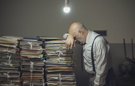 Desperate businessman with lots of paperwork in his messy office at night, he is leaning on a pile of files, bureaucracy and deadlines conceptの写真素材